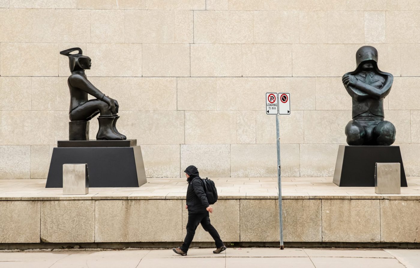 A person walks briskly past the Winnipeg Art Gallery-Qaumajuq, in front of two sculptures by Ivan Eyre, 