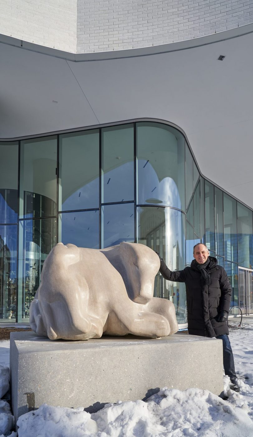 A man poses next to a white Indiana limestone carving outside of the Winnipeg Art Gallery-Qaumajuq that depicts a family of bears climbing over each other.